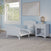The Child Craft Hampton Toddler Bed in Matte White, featuring white bedding and guard rails, sits low beside a matching nightstand in a light blue room. A large window, abstract art, and a potted plant enhance the cozy, modern vibe.