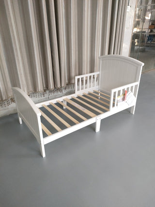 The Child Craft Hampton Toddler Bed in Matte White is a small wooden bed with a slatted base, low-to-the-ground design, and single guard rail, shown on a gray floor in a room with tall curtains and glass walls.