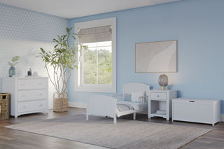 A tidy, light blue children’s bedroom features the Child Craft Hampton Toddler Bed in matte white with guard rails, plus a nightstand, dresser, storage bench, large window for natural light, a plant in a woven basket, and neutral decor accents.