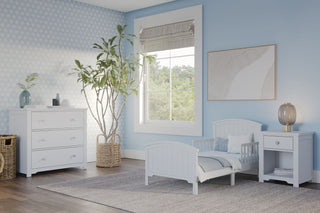 A light blue toddler bedroom includes the Child Craft Hampton Toddler Bed in matte white with guard rails, plus a dresser and nightstand. A large window, tall plant, wall art, and a soft gray rug complete the charming space.