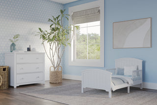 A cozy toddler bedroom features light blue walls, a Child Craft Hampton Toddler Bed in matte white with guard rails, a white dresser, large window, tall potted plant, gray rug, and modern abstract art. Natural light fills the room.