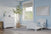 A cozy toddler bedroom features light blue walls, a Child Craft Hampton Toddler Bed in matte white with guard rails, a white dresser, large window, tall potted plant, gray rug, and modern abstract art. Natural light fills the room.