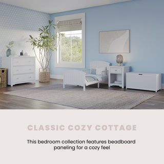 A cozy bedroom with light blue walls, white beadboard furniture including the Child Craft Hampton Toddler Bed in matte white with guard rails, a dresser, nightstand, storage bench, large window, plant, and a soft gray rug. Text: Classic Cozy Cottage.
