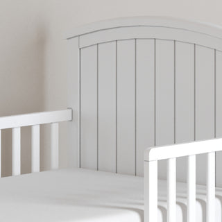 A close-up of the Child Craft Hampton Toddler Bed in Matte White shows vertical slats, an arched headboard, and secure guard rails, ready for use as a toddler bed against a light-colored wall.