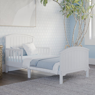 The Child Craft Hampton Toddler Bed in matte white, with guard rails and blue and white bedding, is set on a gray rug in a bright room featuring light blue patterned wallpaper, a leafy plant, and framed wall art above the bed.