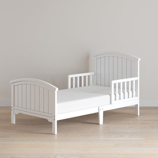 The Child Craft Hampton Toddler Bed in Matte White, featuring vertical slats, sturdy guard rails, and a plain white crib mattress, is set on a light wood floor against a beige wall.