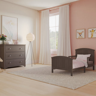 A cozy toddler bedroom with pink walls, a Child Craft Hampton Toddler Bed in Dapper Gray with guard rails and pink bedding, matching dresser, framed animal prints, floor lamp, large window with sheer curtains, and a light gray rug on wood floors.