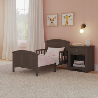 A cozy children’s bedroom with the Child Craft Hampton Toddler Bed in Dapper Gray featuring guard rails, a comfy crib mattress, pink bedding, matching nightstand, wall lamp, animal art, pink polka-dot walls, white curtains, and a sunlit light carpet.