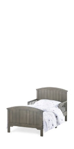 The Child Craft Hampton Toddler Bed in Dapper Gray features a slatted headboard and footboard, fits a standard crib mattress, and is shown with a white and gray patterned comforter and pillow.