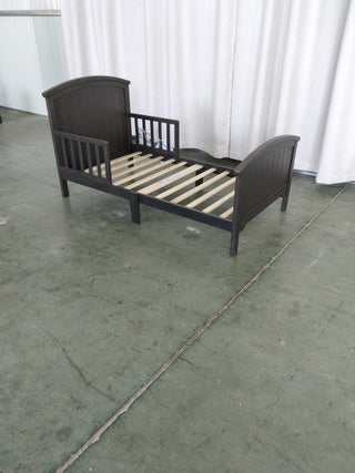 The Child Craft Hampton Toddler Bed in Dapper Gray, featuring slats and raised headboard and footboard, is shown without a crib mattress on a green concrete floor in front of white curtains.