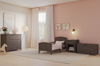 A cozy toddler bedroom with light pink walls, a Child Craft Hampton Toddler Bed in Dapper Gray featuring guard rails, plus a dresser, nightstand, toy chest, and rocking horse. White-curtained window and animal prints complete the décor.