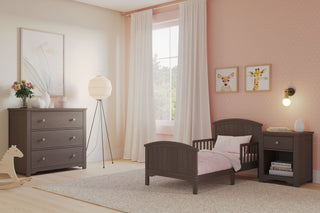 A cozy, pastel child's bedroom with a Child Craft Hampton Toddler Bed in Dapper Gray featuring guard rails, nightstand, dresser; light pink polka dot walls, animal art, white-curtained window, and a gray rug on wood floors.