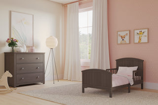 A cozy toddler bedroom with a Child Craft Hampton Toddler Bed in Dapper Gray featuring guard rails, a matching dresser, pink polka dot accent wall, animal art, light curtains, a rocking horse, and soft natural window light.
