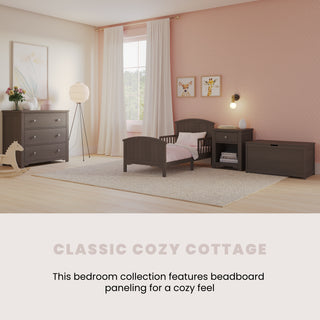 A cozy bedroom featuring the Child Craft Hampton Toddler Bed in Dapper Gray with a crib mattress and guard rails, dresser, nightstand, toy chest, rocking horse, pink polka dot wallpaper, sheer curtains, and framed artwork. Classic Cozy Cottage.