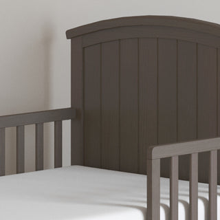 A close-up view of the Child Craft Hampton Toddler Bed in Dapper Gray with sturdy guard rails and a white crib mattress, set against a light-colored wall.