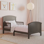 The Child Craft Hampton Toddler Bed in Dapper Gray with guard rails sits in a softly lit room with light pink bedding, pink polka dot walls, animal art, a floor lamp, and light curtains.