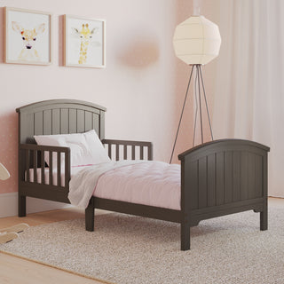 The Child Craft Hampton Toddler Bed in Dapper Gray with guard rails sits in a softly lit room with light pink bedding, pink polka dot walls, animal art, a floor lamp, and light curtains.