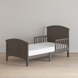 The Child Craft Hampton Toddler Bed in Dapper Gray, featuring vertical slat headboard and footboard, low guard rails, and a white crib mattress, is placed on a light wood floor against a plain beige wall.