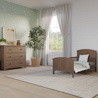 A cozy nursery features the Child Craft Hampton Toddler Bed in Dusty Heather, a matching dresser, soft area rug, a potted plant, green star-patterned wallpaper on one wall, and white curtains framing a large window.