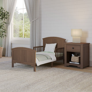 The Child Craft Hampton Toddler Bed in Dusty Heather stands beside a matching nightstand with a lamp, set against a white shiplap wall in a bright room featuring a large window, curtains, a plant, and a gray rug.