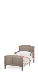The Child Craft Hampton Toddler Bed in Dusty Heather features a wooden frame with headboard and footboard, both with vertical slats. It's shown with white bedding and a pink-green floral comforter against a plain white background.