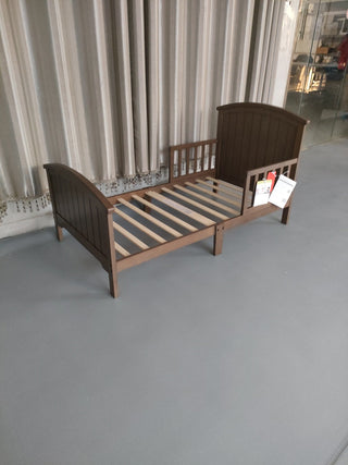 The Child Craft Hampton Toddler Bed in Dusty Heather features a slatted base, arched headboard and footboard, and stands on a gray floor near beige curtains, with price tags attached to the side.