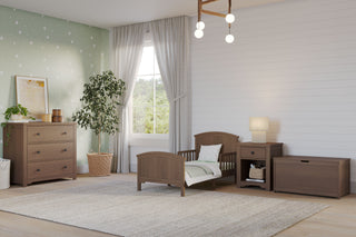A cozy, light-filled children’s bedroom features the Child Craft Hampton Toddler Bed in Dapper Gray, plus wood dresser, nightstand, toy chest, green star-patterned wall, large rug, potted plant, and window with sheer curtains.