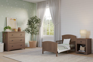 A cozy children's bedroom features the Child Craft Hampton Toddler Bed in Dusty Heather, a nightstand, dresser, green star-patterned accent wall, large window with white curtains, potted tree, beige rug, and light wood flooring.