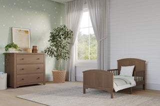 A cozy children’s bedroom features the Child Craft Hampton Toddler Bed in Dusty Heather, a matching dresser, soft rug, potted plant, light curtains, and star-patterned green wallpaper. Sunlight pours through a large window.