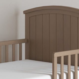 A close-up view of the Child Craft Hampton Toddler Bed in Dusty Heather, showcasing its curved headboard, vertical slats, and a white mattress.
