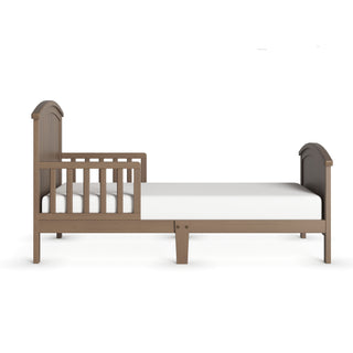 The Child Craft Hampton Toddler Bed in Dusty Heather features a sturdy wood frame, partial safety rail, curved headboard and footboard with a light brown finish, and comes with a white mattress for simple, safe comfort.