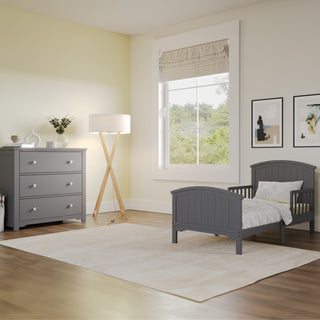 A bright, modern kids’ room with the Child Craft Hampton Toddler Bed in Cool Gray with guard rails, a matching dresser, standing lamp, light rug, framed art on pale yellow walls, and a window overlooking greenery.