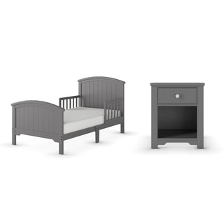 The Child Craft Hampton Toddler Bed in Cool Gray, featuring a low profile, sturdy guard rails, and arched headboard, is shown beside a matching gray nightstand with one drawer and an open shelf underneath.
