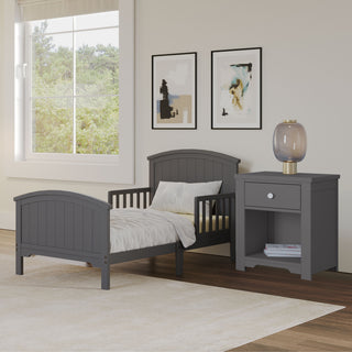 A modern toddler bedroom features a gray bed, white bedding, and the Child Craft Harmony Nightstand in Cool Gray with bedside storage and child-safe finishes. Abstract art decorates the walls, while a large window fills the room with natural light.