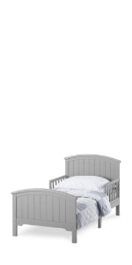 The Child Craft Hampton Toddler Bed in Cool Gray features a slatted headboard and footboard, sits low to the ground for easy access, and is shown with a light blue comforter and matching pillow.