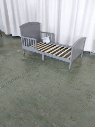 The Child Craft Hampton Toddler Bed in Cool Gray, featuring a slatted base and curved headboard and footboard, stands low on a green concrete floor before white curtains. A paper sign rests on its slats.