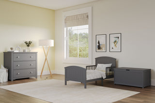 A bright, modern child's room features the Child Craft Hampton Toddler Bed in Cool Gray with guard rails, a matching dresser, and a storage bench. Light walls, a large window with Roman shade, framed art, floor lamp, and a light rug complete the cozy space.