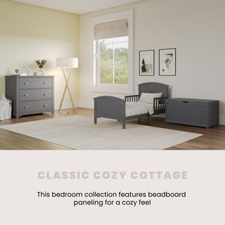 A cozy bedroom featuring the Child Craft Hampton Toddler Bed in Cool Gray with guard rails, paired with matching beadboard chest and dresser. Light walls, a floor lamp, wall art, and a big window add warmth and charm.