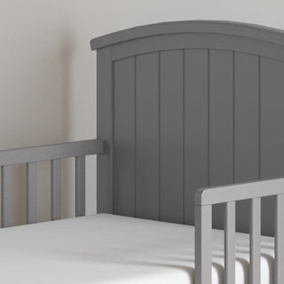 The Child Craft Hampton Toddler Bed in Cool Gray features vertical slats, a curved headboard, and guard rails. Shown with a white mattress, this stylish bed is pictured in a softly lit room with a plain wall background.