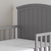 The Child Craft Hampton Toddler Bed in Cool Gray features vertical slats, a curved headboard, and guard rails. Shown with a white mattress, this stylish bed is pictured in a softly lit room with a plain wall background.
