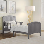 The Child Craft Hampton Toddler Bed in Cool Gray sits low to the ground with guard rails in a softly lit bedroom, styled with a white comforter, wooden floor, beige rug, standing lamp, abstract wall art, and a right-side window.