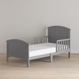 The Child Craft Hampton Toddler Bed in Cool Gray features guard rails, paneled headboard and footboard, and a white mattress, shown on light wooden flooring against a beige wall.