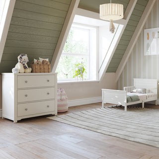 Bright attic nursery with green sloped ceilings, window, soft natural light, small white toddler bed, striped rug, and Child Craft Harmony 3-Drawer Dresser with Changing Table Topper in Brushed Cotton holding toys.
