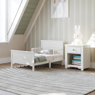 A Child Craft Harmony Nightstand in Brushed Cotton sits beside a small white bed with a bunny lamp in a cozy attic room featuring striped wallpaper, framed art, and a circular striped rug on the floor.