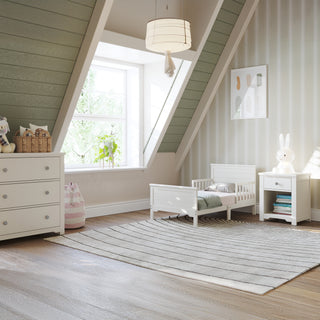 A cozy attic nursery with slanted ceilings features a toddler bed, nightstand, Child Craft Harmony 3-Drawer Dresser with Changing Table Topper in Brushed Cotton, stuffed toys, striped rug, and a large window for natural light. Soft walls and a framed print complete the space.