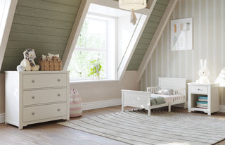 A cozy kids bedroom with slanted green walls, a small white bed, a Child Craft Harmony 3-Drawer Dresser with Changing Table Topper in Brushed Cotton, toys, nightstand with books, striped rug, sunlight, and a plant on the windowsill.