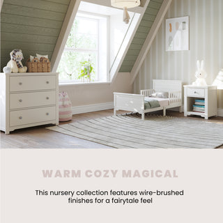A cozy nursery with the Child Craft Woodland Toddler Bed in Brushed Cotton, matching dresser, nightstand, stuffed animals, and pastel decor under slanted windows. Wire-brushed finishes create a warm, magical, fairytale feel. Text reads: WARM COZY MAGICAL.