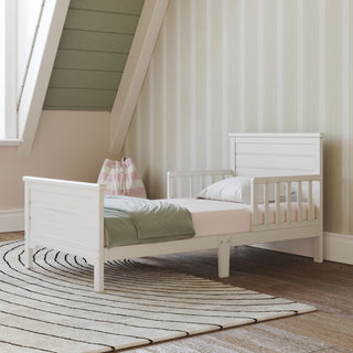 The Child Craft Woodland Toddler Bed in brushed cotton, featuring guardrails, is placed on a circular-patterned rug in a cozy room with striped wallpaper and a wooden floor. A pink and green throw and pillow adorn the bed, with a fabric bag beside it.
