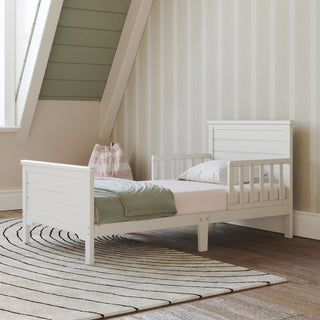 The Child Craft Woodland Toddler Bed in brushed cotton with side rails is dressed in pale pink bedding and a green blanket. It stands on a striped circular rug in a cozy room with striped wallpaper, a slanted ceiling, and a pink-striped bag by the bed.