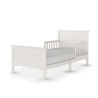 The Child Craft Woodland Toddler Bed in Brushed Cotton features a modern farmhouse style with slatted headboard and footboard, low profile, partial toddler bed rails, and is shown with a plain mattress.